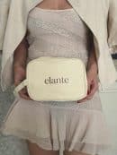 A close-up view of a woman, dressed in a beige linen jacket and a matching sparkling sequined ruffled mini dress, holding a beige canvas Elante branded rectangular pouch bag with both hands. The bag features the black text "elante" in a sans-serif font.